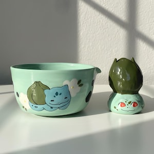 May include: A mint green ceramic bowl and matching decorative bud vase. The bowl features a hand-painted Bulbasaur design with white flowers and black spots. The vase is a Bulbasaur head with red eyes.