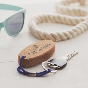 Op de afbeelding: Een kurken sleutelhanger met het woord "tartaruga" erop gedrukt, een blauw koord en een set sleutels. De sleutelhanger ligt naast een mintgroene zonnebril en een wit touw. De sleutelhanger is ongeveer 7.6 cm lang.