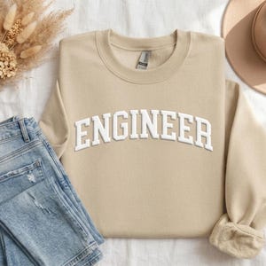 May include: A beige sweatshirt displaying the word "ENGINEER" in white, block lettering. The garment is folded, with blue jeans and a beige hat in view. The sweatshirt features a crew neck and long sleeves.