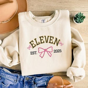 Op de afbeelding: Crèmekleurige sweatshirt met het woord "ELEVEN" in gouden letters, een roze strik en het jaar "2025". De sweatshirt is opgevouwen op een houten oppervlak, met een spijkerbroek en een bruine hoed zichtbaar.