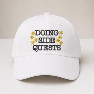 Könnte beinhalten: Eine weiße Baseballkappe mit schwarzem Sticktext, der DOING SIDE QUESTS lautet. Der Text wird auf beiden Seiten von kleinen, gelben, sechsstrahligen sternartigen Symbolen flankiert, die vertikal angeordnet sind. Die Kappe hat eine feste Krone.