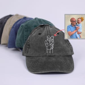 Op de afbeelding: Een rij van vijf verweerde baseballpetten in verschillende kleuren: houtskool, beige, marineblauw, groen en donkergrijs. De voorste pet heeft een witte geborduurde omtrek van een persoon die een kind vasthoudt. Een kleine foto toont een man die een baby vasthoudt.