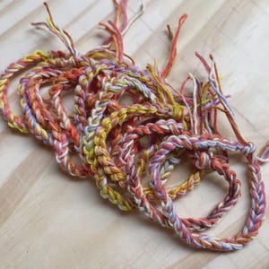 May include: Braided bracelets in a variety of colors, including yellow, orange, pink, and white. The bracelets are made of twisted yarn and are arranged on a light wooden surface. The ends of the yarn are visible, adding to the handmade aesthetic.