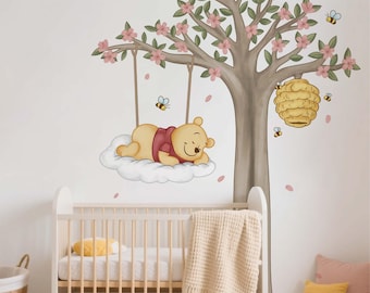 Puede incluir: Un vinilo decorativo para la pared de una guardería con un oso de dibujos animados durmiendo en un columpio de nube bajo un árbol con flores rosas. Una cuna con un cojín amarillo y una manta beige está en primer plano. También se representan abejas y una colmena.