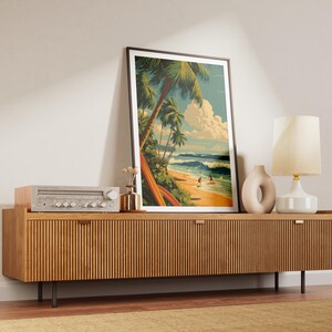 May include: A framed art print depicting a tropical beach scene with palm trees, surfers, and waves. The artwork is displayed on a wooden cabinet with a vintage stereo, a vase, and a lamp.