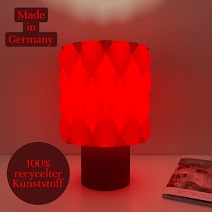Könnte beinhalten: Eine rote Tischlampe mit geometrischem Muster und zylindrischem Sockel. Die Lampe leuchtet und wirft einen roten Schein. Auf der Lampe steht "Made in Germany" und "100% recycled Kunststoff". Ein Buch ist auf dem Tisch zu sehen.