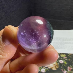 May include: A translucent, spherical crystal ball with a swirling pattern of purple, pink, and green hues. The polished sphere is held in a hand, with a dark background and a printed page visible.