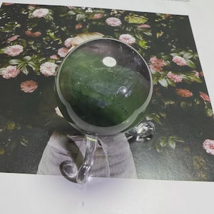 May include: A green and purple crystal ball on a clear stand, set against a backdrop of pink flowers. The crystal ball has a smooth, polished surface and reflects the light. The stand is made of clear acrylic.