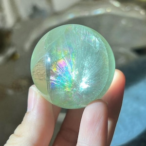 May include: A translucent, light green crystal sphere. The sphere has internal rainbow-colored inclusions that catch the light. The crystal is held in a person's hand, with the background out of focus.