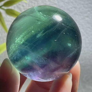 May include: A translucent crystal ball with a blend of green, blue, and purple hues. The sphere is held in a hand, with the colors shifting and blending throughout the polished surface. The crystal ball is a decorative item.