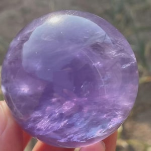 May include: A polished amethyst crystal sphere, showcasing a range of purple hues. The sphere is held in a hand, with the background blurred, emphasizing the crystal's smooth, reflective surface. The crystal's color varies from light lavender to deep violet.
