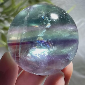 May include: A clear, spherical fluorite crystal ball with bands of purple, green, and iridescent colors. The polished gemstone is held in a hand, showcasing its smooth surface and natural beauty. A decorative item.