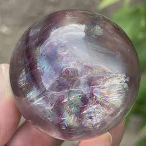May include: A clear, spherical crystal ball with a purple and iridescent sheen. The crystal reflects light, creating a rainbow effect across its surface. The sphere is held in a hand, with a blurred green background.
