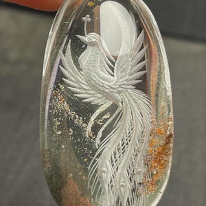 May include: A clear, oval-shaped pendant with a detailed, white phoenix carving. The bird is set against a backdrop of a white moon and earthy inclusions. The pendant is held by a hand.