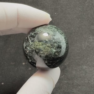 May include: A polished spherical stone featuring a deep dark green and black base colour with mottled grey and metallic gold-toned mineral inclusions throughout the surface, reflecting light with a smooth, glossy, and reflective finish.
