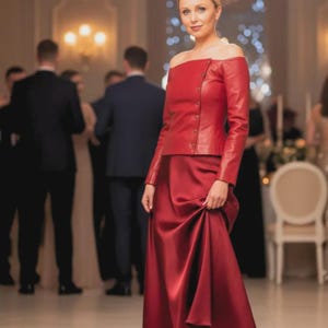 Pode incluir: Um conjunto vermelho formal com uma saia longa e fluida de cetim e um top de couro a condizer com os ombros descobertos e detalhes de botões. A roupa é usada numa sala bem iluminada com outras pessoas ao fundo.