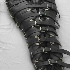 Op de afbeelding: Een zwarte, lederachtige full-body fixatiezak voorzien van een reeks horizontale banden met zilverkleurige metalen gespen en D-ringen over de gehele lengte, inclusief een bijpassende hoofdbedekking met een gesp bij de nek.