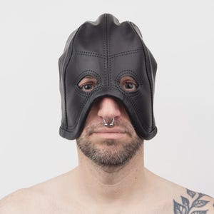 Könnte beinhalten: Eine schwarze Lederhaube mit verstärkten Nähten und zwei kreisförmigen Augenöffnungen. Die Haube bedeckt den oberen Teil des Kopfes und die Stirn, während Nase, Mund und Kinn frei bleiben. Die Konstruktion weist sichtbare schwarze Nähte auf.