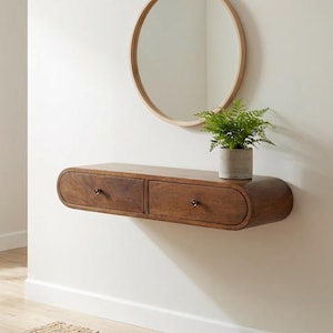 May include: A floating wooden console table with two drawers, a round mirror, and a small potted plant. The console is mounted on a white wall above a woven jute rug. The mirror has a light wood frame.