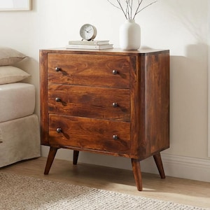 Op de afbeelding: Houten dressoir met drie lades en een warme bruine afwerking. Het dressoir heeft een mid-century modern design met taps toelopende poten en ronde knoppen. Een witte vaas met takken, een klok en boeken staan erop. In een slaapkamer.