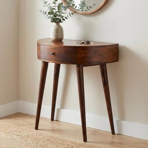 May include: A dark brown, semi-circular wooden console table with a single drawer and three angled legs. A small vase with greenery sits on top, along with keys. A round mirror hangs on the wall above the table.