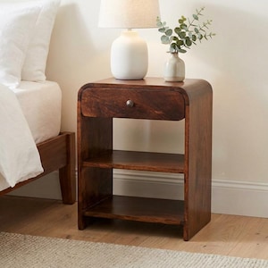 May include: A dark brown wooden bedside table with a curved top edge, one drawer featuring a small round knob, and two open storage shelves. A white ceramic table lamp with a beige fabric shade and a small vase with green leaves sit on top.