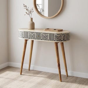 May include: A light wood console table with a black and white geometric pattern on the front. The table has four angled legs and a small, round mirror above it. A vase with dried flowers sits on the table.