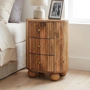 May include: A wooden bedside table with three drawers and a ribbed design. The table has a round shape and sits on three rounded feet. A lamp and a framed photo are on top. The table is next to a bed with beige bedding. The table is approximately 60 cm high.