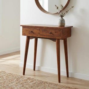 May include: A brown wooden console table featuring two drawers with small black knobs, supported by four tapered legs. A small ceramic vase containing dried eucalyptus stems sits on the tabletop surface, positioned below a round wall mirror.