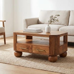 May include: A wooden coffee table with two drawers and a lower shelf. The table has a warm brown finish, brass drawer pulls, and rounded legs. A vase with greenery and two books sit on top. The table is 48 inches long, 24 inches wide, and 18 inches high.