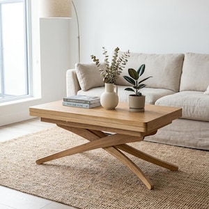 May include: A rectangular coffee table made of light wood, featuring a cross-legged base. The table holds a vase with dried eucalyptus, a potted plant, and two books. The table is placed on a jute rug in a living room setting.