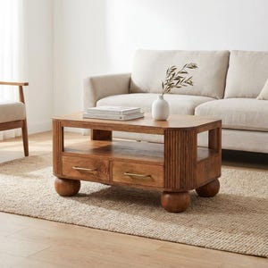 Op de afbeelding: Een houten salontafel met twee lades en messing handgrepen. De tafel heeft een onderste plank en wordt ondersteund door vier afgeronde poten. Een vaas met groen en twee boeken staan op de tafel.