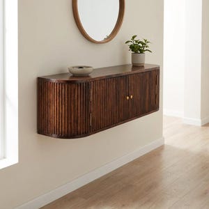 May include: A wall-mounted dark brown wooden console cabinet featuring a curved, fluted design with vertical slats, two central doors with small gold-toned knobs, and rounded side edges. A small gray bowl and a green potted plant sit on top.
