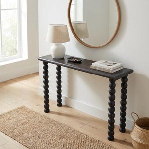 May include: A dark brown console table with four legs featuring a stacked ball design. The table surface holds a white lamp with a beige shade, a small black tray with keys, and two stacked books. A round mirror hangs on the wall above the table.