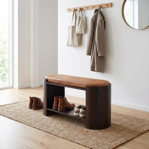 Può includere: Panca in legno marrone scuro con seduta in pelle marrone. La panca ha un ripiano inferiore con scarpe e stivali. Un tappeto di iuta è sotto la panca. Un attaccapanni con un cappotto e borse appese al muro.