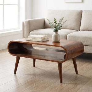 Peut inclure: Table basse en bois brun foncé de style moderne du milieu du siècle. Elle présente une forme incurvée, une étagère inférieure et des pieds effilés. Un vase avec de la verdure et des livres sont dessus. Mesure environ 120 cm de long.