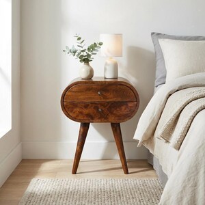 Peut inclure: Une table de chevet en bois brun foncé de forme ovale, dotée de deux tiroirs avec de petits boutons ronds et de quatre pieds fuselés. Une lampe de table en céramique blanche et un petit vase blanc contenant des tiges d'eucalyptus vert.
