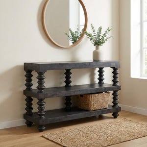 Può includere: Una consolle in legno grigio scuro a tre ripiani con gambe tornite. Un cestino intrecciato è posto sul ripiano inferiore. Uno specchio rotondo è appeso alla parete sopra il tavolo e un vaso con verde è sul ripiano superiore.