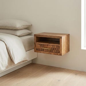 Könnte beinhalten: Ein schwebender Nachttisch aus hellem Holz mit Schublade und offenem Fach. Die Schublade hat ein geometrisches Muster und einen goldfarbenen Griff. Der Nachttisch ist an einer beigefarbenen Wand befestigt. Maße ca. 40 cm x 30 cm x 20 cm.