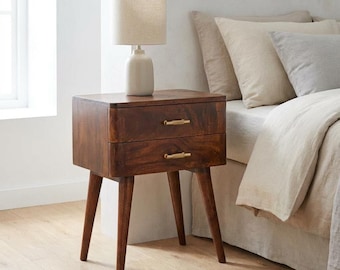 May include: A dark brown wooden bedside table with two drawers, each featuring a horizontal brass-toned metal handle. The bedside table stands on four tapered legs and supports a table lamp with a cream-coloured cylindrical fabric shade and base.