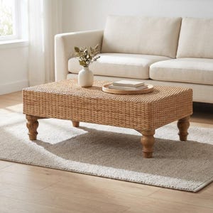 May include: A rectangular coffee table featuring a woven light brown rattan surface and four turned wooden legs. On top sits a round wooden tray holding two stacked books and a white ceramic vase containing dried botanical stems.