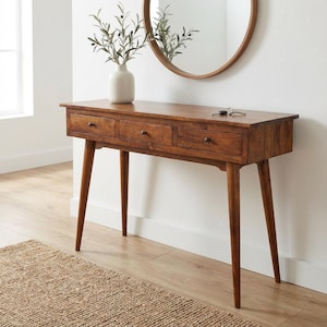 May include: A mid-century modern style wooden console table with three drawers and tapered legs. The table is a warm brown color and has a natural wood grain. A vase with greenery and a round mirror are on the wall behind the table.