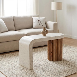 May include: A modern coffee table with a curved, speckled white top and a cylindrical base made of vertical wooden slats. A beige vase with a twig sits on the table. The table is placed on a woven rug.