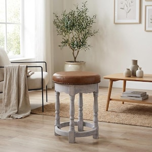Puede incluir: Un taburete redondo con asiento de cuero marrón y base de madera gris claro envejecida. El taburete tiene un borde festoneado y patas torneadas, lo que le da un aspecto vintage. El taburete mide aproximadamente 46 cm de diámetro y 46 cm de altura.