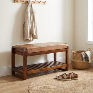 May include: A wooden entryway bench with a brown leather seat and a slatted lower shelf. A pair of brown and tan sneakers sits on a round jute rug. A woven basket and a coat rack with a beige jacket complete the scene.