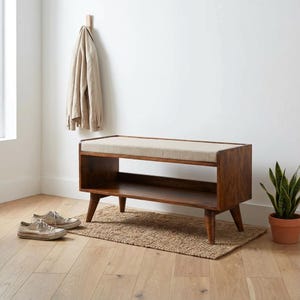 Op de afbeelding: Een rechthoekige houten bank met een donkerbruine afwerking, voorzien van een beige gestoffeerd zitkussen aan de bovenkant en een open opbergplank eronder. De bank rust op vier taps toelopende poten en staat op een geweven rechthoekig kleed.
