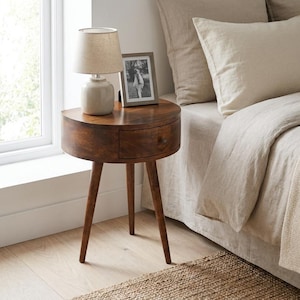 Può includere: Un comodino in legno a forma di semicerchio con un cassetto singolo e tre gambe affusolate. Una piccola lampada con paralume in tessuto beige e una fotografia con cornice grigia poggiano sulla superficie piatta del tavolo, posizionato accanto a un letto.