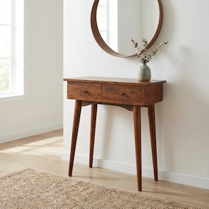 May include: A brown wooden console table featuring two drawers with small black knobs and four tapered legs. A round wooden-framed mirror is mounted on the wall above the table, and a small ceramic vase with dried eucalyptus sits on top.