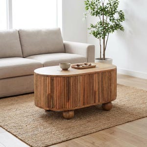 May include: An oval wooden coffee table with a ribbed design and rounded legs. The table is light brown and sits on a woven jute rug. A beige sofa is in the background.