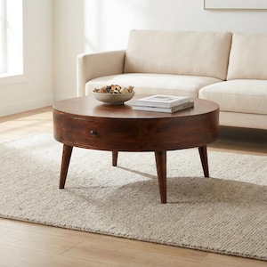 May include: A round, dark brown wooden coffee table featuring a single front-facing drawer with a small, dark circular knob. The table is supported by four tapered legs and holds a shallow bowl filled with decorative items and two stacked books.